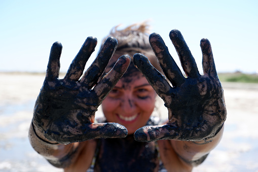 Woman who doing mud therapy shows her hands covered with healing sulfurous black mud. Her smiling face appearing from hands blurred in background.