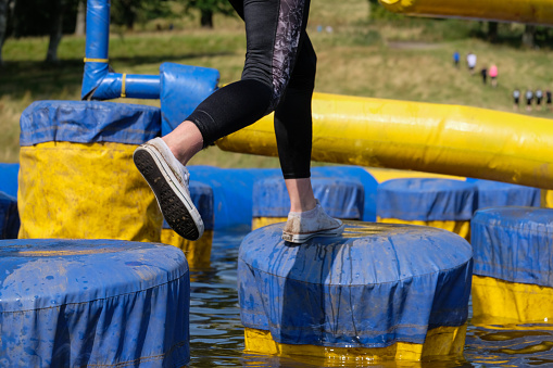 A woman jumping across a blue and yellow obstacle course, leaping from point to point, as large inflatable arms try and knock her into the water below. She is wearing black leggings and white coloured sports shoes. She is trying to catch up her team mates who are running ahead and can be seen in the background running up a grassy hill