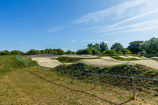 A bmx dirt track surrounded by metal fence, grass and trees under a majestic blue sky and some white clouds