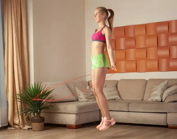 young blonde woman in sportswear jumping with jumping rope at home, cardio training in living room