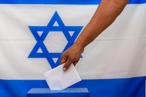 Israeli man putting a ballot in a ballot box on election day. Close up of hand with white votes paper on Israel flag background. Mockup, space for text.