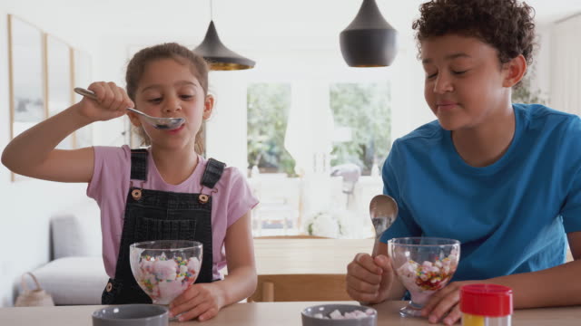 Children In Kitchen At Home Eating Ice Cream And Pulling Faces At Each Other