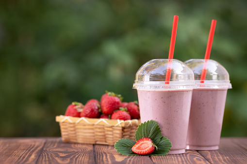 two disposable glasses of strawberry milkshake or smoothie and fresh ripe berries in basket on wooden table outdoors