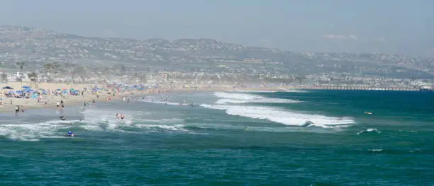 Crowd of beachgoers and strong surf at Newport Beach with Balboa Pier and Laguna hils in the background