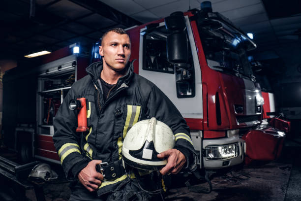 istock Fireman wearing uniform holding a helmet and looking sideways while standing near a fire truck in a garage of a fire department 1171678197