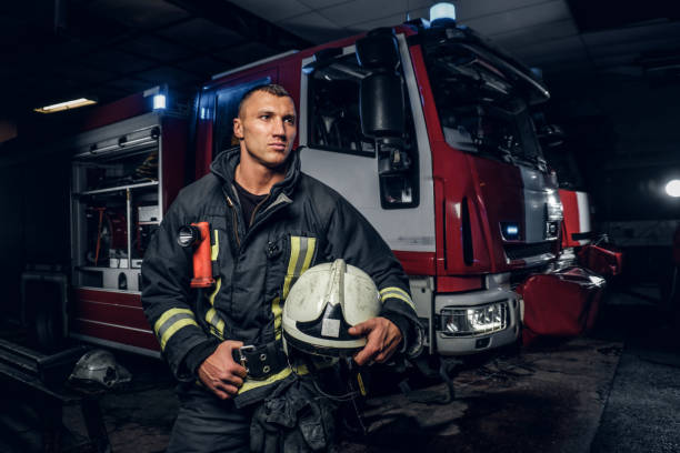 istock Fireman wearing uniform holding a helmet and looking sideways while standing near a fire truck in a garage of a fire department 1171679525