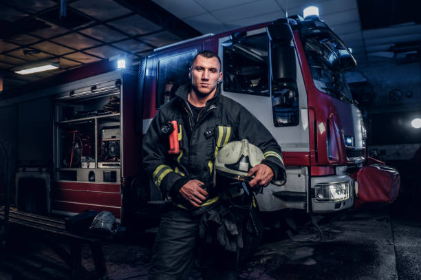 istock Fireman wearing uniform holding a helmet and looking at a camera while standing near a fire truck in a garage of a fire department 1171679736