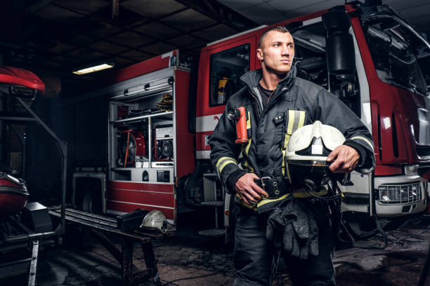 istock Fireman wearing uniform holding a helmet and looking sideways while standing near a fire truck in a garage of a fire department 1171679966