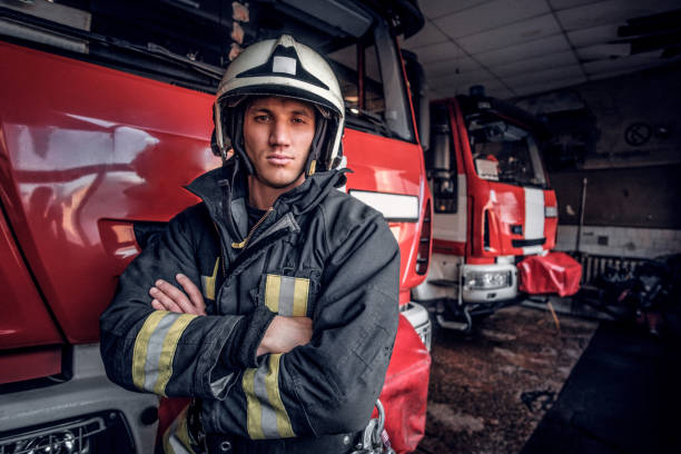 A confident fireman wearing protective uniform standing next to a fire engine in a garage of a fire department, crossed arms and looking at a camera stock photo