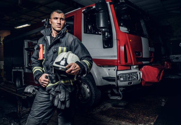 Fireman wearing uniform holding a helmet and looking sideways while standing near a fire truck in a garage of a fire department
