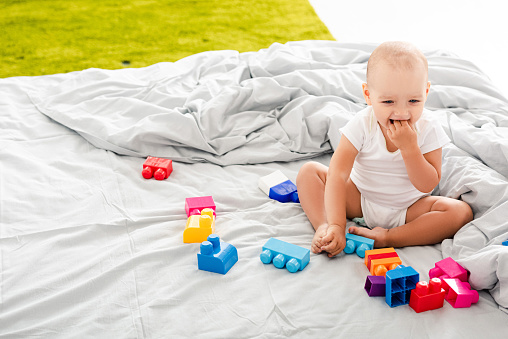 Funny barefoot baby in white clothes sitting on bed near colorful construction and taking fingers into his mouth