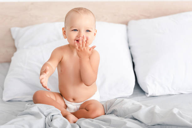adorabile bambino scalzo in pannolino sorridente mentre è seduto su biancheria da letto bianca - one baby girl only foto e immagini stock