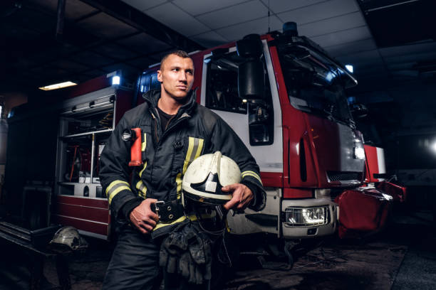 istock Fireman wearing uniform holding a helmet and looking sideways while standing near a fire truck in a garage of a fire department 1171680702