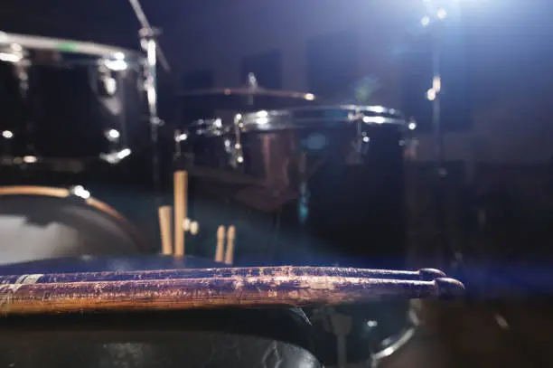 Photo of Close-up of used drumsticks lie on a drummer's chair against the background of a drum kit and flare of spotlights.