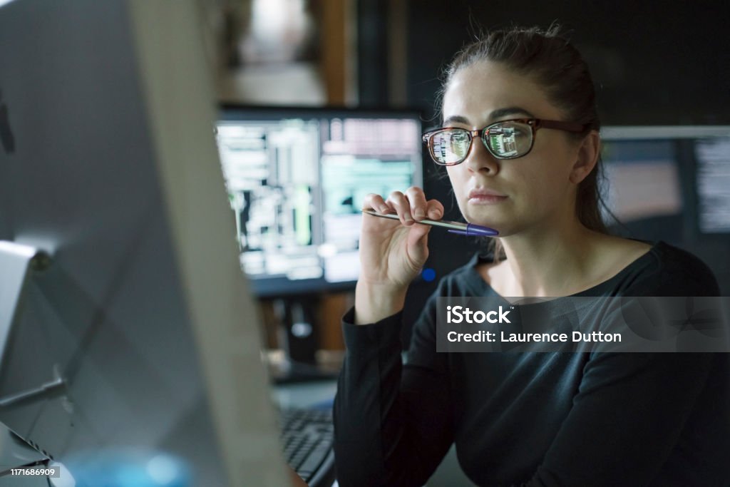 Woman monitors dark office Stock photo of young woman’s face as she contemplates one of the many computer monitors that surround her. Data Stock Photo