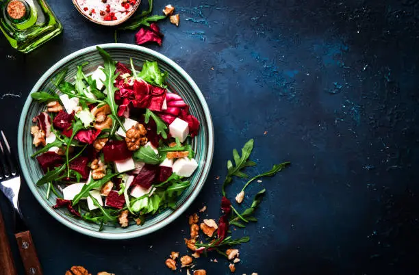 Photo of Arugula, Beet and cheese salad with fresh radicchio and walnuts on plate with fork, dressing and spices on blue kitchen table background, place for text, top view