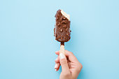 Young woman hand holding white vanilla ice cream with nuts and chocolate glaze on pastel light blue background. Bitten food. Closeup. Point of view shot. Top view.