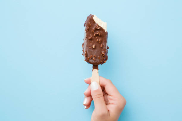 Young woman hand holding white vanilla ice cream with nuts and chocolate glaze on pastel light blue background. Bitten food. Closeup. Point of view shot. Top view. Young woman hand holding white vanilla ice cream with nuts and chocolate glaze on pastel light blue background. Bitten food. Closeup. Point of view shot. Top view. ice cream photos stock pictures, royalty-free photos & images