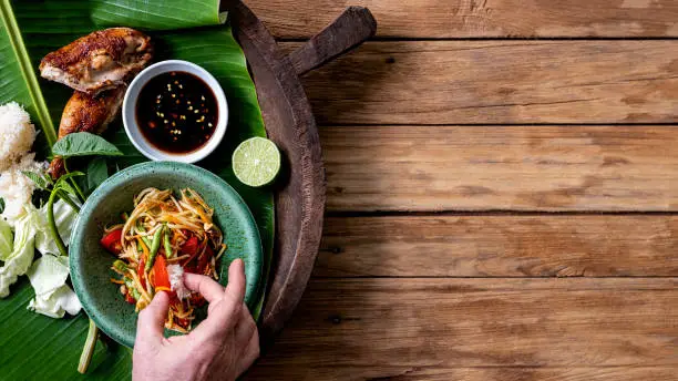 Western man eating traditionally with his hands, following Thai local culture the indigenous fresh world famous Som Tam (papaya salad) with BBQ chicken, sticky rice and raw salad vegetables on an old wooden table background.  Good copy space to the right of the image.
