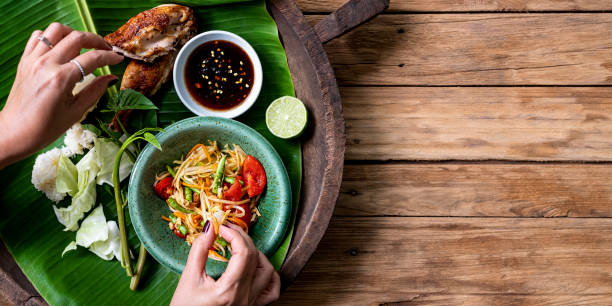 señora tailandesa comiendo tradicionalmente con sus manos, fresco mundialmente famoso som tam (ensalada de papaya) con pollo barbacoa, arroz pegajoso y verduras crudas de ensalada sobre un viejo fondo de mesa de madera. - asian cuisine fotografías e imágenes de stock