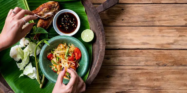 Thai lady eating traditionally with her hands, fresh world famous Som Tam (papaya salad) with BBQ chicken, sticky rice and raw salad vegetables on an old wooden table background.  Good copy space to the right of the image.