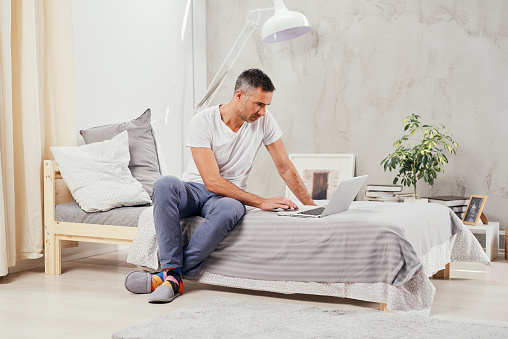 Handsome Caucasian middle aged man dressed casual using laptop while sitting on bed in bedroom.
