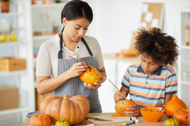 Concentrated young black mother in apron creating Halloween decorations with son in workshop Concentrated young black mother in apron creating Halloween decorations with son in workshop pumpkin decorating stock pictures, royalty-free photos & images