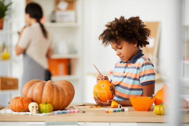istock Curious talented boy with Afro hairstyle standing at counter with art and craft tools and painting pumpkin with paintbrush 1171715482