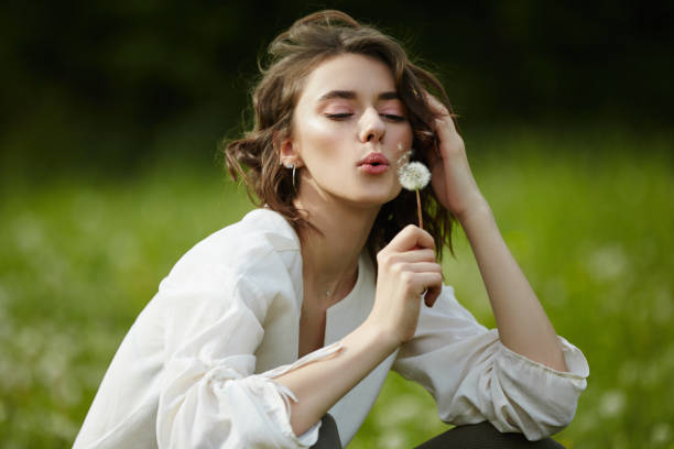 spring portrait of a girl sitting in a field on the grass among dandelion flowers. cheerful girl enjoys sunny spring weather. natural beauty of a woman, natural cosmetics - nature beauty women fashion model imagens e fotografias de stock