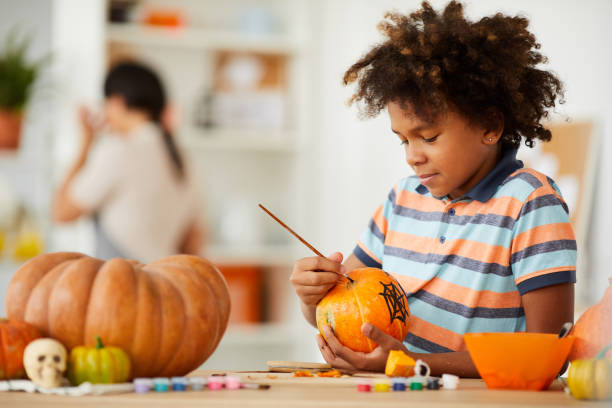Smiling creative child with Afro hairstyle leaning on counter with gouaches and making design on pumpkin
