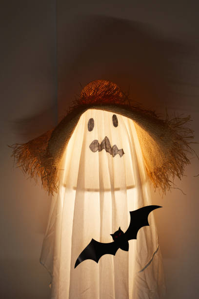 istock Illuminated lamp covered with sheet placed in dark room, white sheet with drawn ugly face and straw hat 1171720059