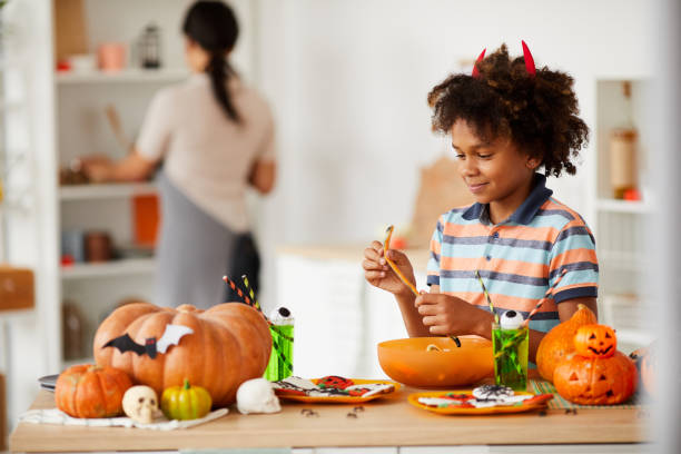 istock Smiling cute black boy with devils horns standing at counter with various Halloween candies and eating marmalade worm 1171720454