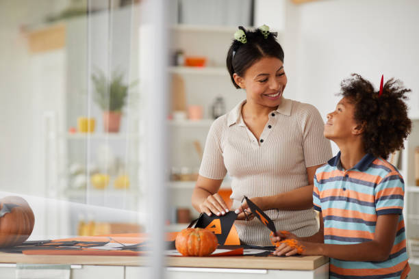 Cheerful young black mother and cute son with Afro hairstyle standing at counter and preparing paper decorations for Halloween with pleasure Cheerful young black mother and cute son with Afro hairstyle standing at counter and preparing paper decorations for Halloween with pleasure carving food photos stock pictures, royalty-free photos & images