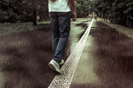 A man in jeans and white t-shirt walking alone in the park alley on a rainy evening