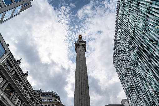 Low angle view of the monument to the great gire of London. Buildings surrounding tower are in view. Sky is with high clouds. No people are seen in frame. Shot with a full frame mirrorless digital camera.