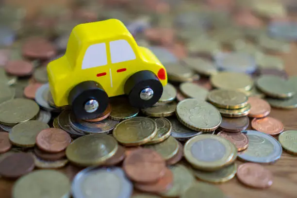 Wooden toy car, yellow in color, over a pile of money, euro coins. Suitable for example for the concept of car purchase or car insurance.