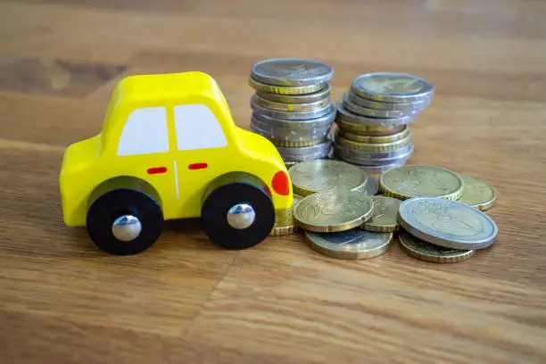 Wooden toy car, yellow in color, next to a pile of euro coins.