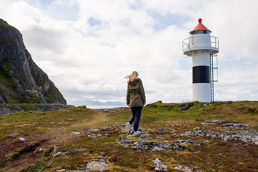 Blond hair woman walks near the lighthouse and enjoy beautiful nature landscape. Lonely girl. Amazing scenic outdoors view in North. Travel and adventure. Explore Norway. Lofoten Islands. Scandinavia