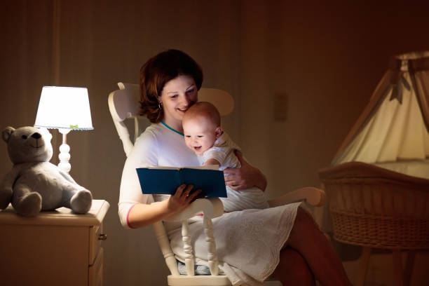 Mother reading a book to little baby Mother and baby reading a book in dark bedroom. Mom and child read books before bed time. Family in the evening. Kids room interior with night lamp and bassinet. Parent holding infant next to crib. crib photos stock pictures, royalty-free photos & images