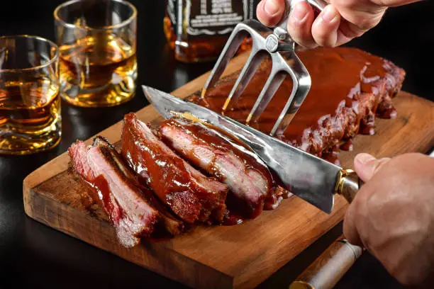 Smoked ribs served with barbecue sauce and whisky cup on dark background. Man hand cutting the ribs.