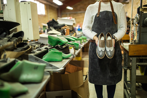 Unrecognizable woman with apron standing in factory and holding in hands model of leather, oxford shoes