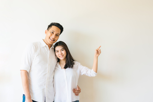 Happy Asian couple in white shirt smiling and pointing finger at empty copy space on the wall