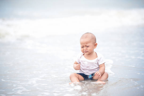 Asian ten month baby girl sitting and crying on the beach, worrying mother will leave her alone. Baby worries playing the beach first time. Asian ten month baby girl sitting and crying on the beach, worrying mother will leave her alone. Baby worries playing the beach first time. outing tantarum in babies stock pictures, royalty-free photos & images