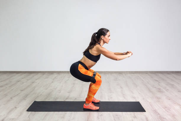 Side view portrait of young sporty healthy beautiful woman in black top and orange leggings doing squatting with elastic resistance band. Side view portrait of young sporty healthy beautiful woman in black top and orange leggings doing squatting with elastic resistance band. Isolated, white wall, indoor, workout concept, looking away crouching stock pictures, royalty-free photos & images