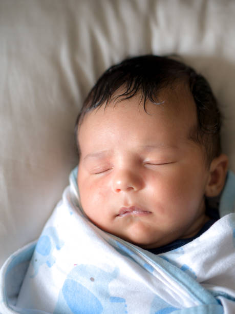 Closeup head shot portrait of a mixed race handsome and cute baby boy with lots of brown hair sleeping and swaddled in a white and blue blanket laying on a bed. stock photo