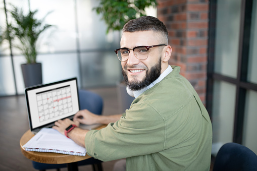 Businessman with laptop. Beaming young businessman in glasses working on laptop and compiling schedule