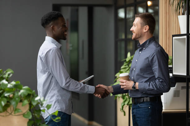Two happy diverse male colleagues handshaking standing in modern office Two happy diverse male colleagues greeting handshaking talking standing in modern office, smiling african and caucasian business men professionals shake hands express respect make deal at meeting office buddies stock pictures, royalty-free photos & images