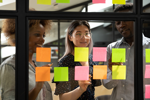 Multiethnic creative diverse young business team people three colleagues students group brainstorm on work plan talk write ideas tasks on post it sticky notes look at glass at scrum teamwork briefing
