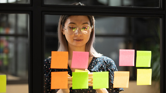 Serious focused young female coach teacher student asian business woman working on project strategy plan writing target tasks creative ideas on sticky post it notes on glass scrum board office wall