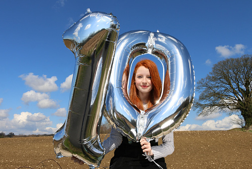 Stock photo showing a beautiful, ginger haired teenager sitting in the countryside on a stonewall holding two silver helium balloons. The balloons form the number ten with one balloon being a number 1 and the other digit a zero.